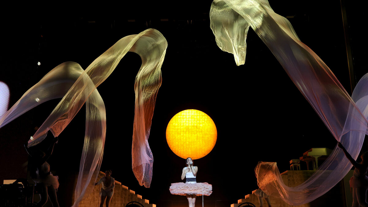 A female singer in a white dress and tutu performs on a stage with a large glowing orange sphere behind her, surrounded by flowing translucent ribbons.