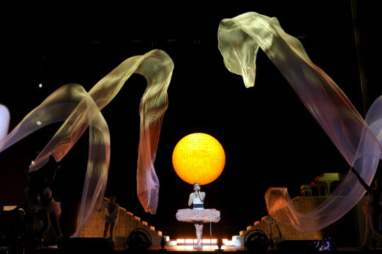 A female singer in a white dress and tutu performs on a stage with a large glowing orange sphere behind her, surrounded by flowing translucent ribbons.