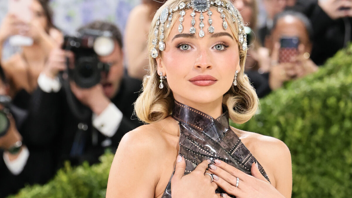 Woman wearing a sparkling jeweled headpiece and metallic halter gown, posing with hands over her chest as photographers look on.