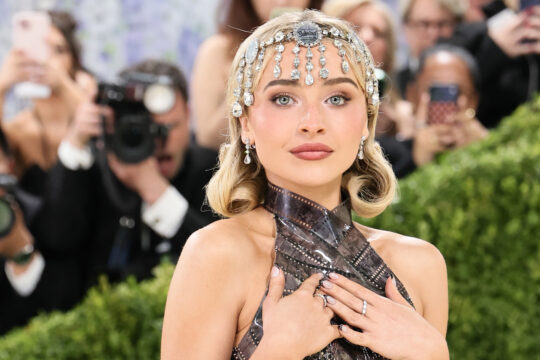 Woman wearing a sparkling jeweled headpiece and metallic halter gown, posing with hands over her chest as photographers look on.