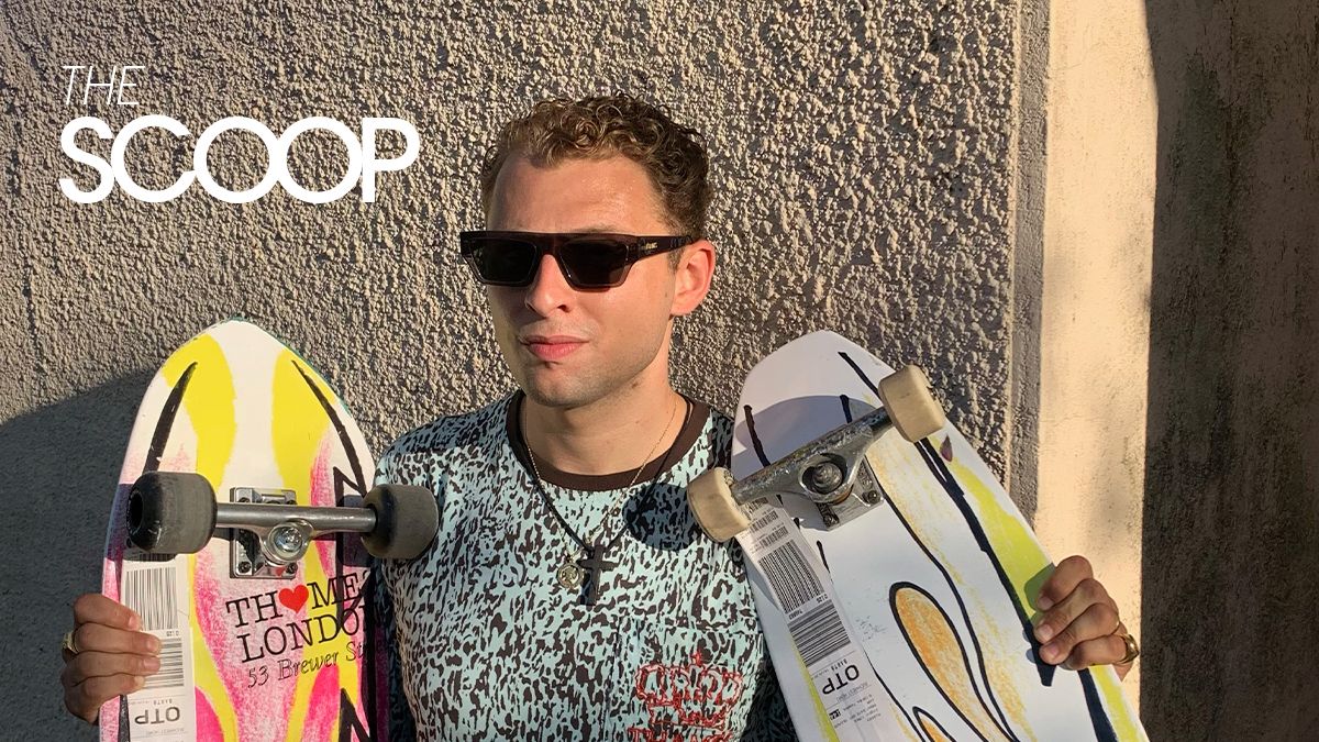 Man wearing sunglasses holds two skateboards beside a textured wall, with 'THE SCOOP' logo in the upper left.