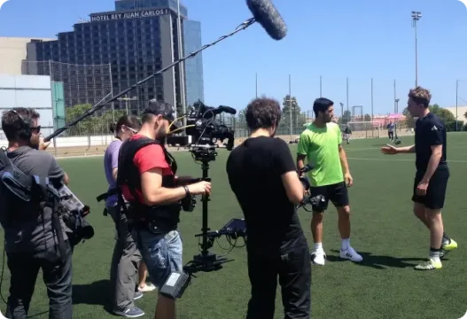 Film crew on a sports field filming two players during an interview; a boom mic hovers above while one player in a green shirt talks to a coach in black.