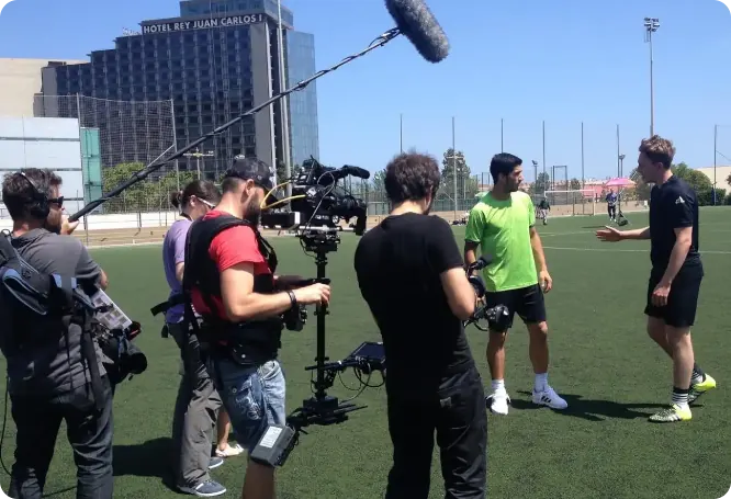 Film crew on a sports field filming two players during an interview; a boom mic hovers above while one player in a green shirt talks to a coach in black.