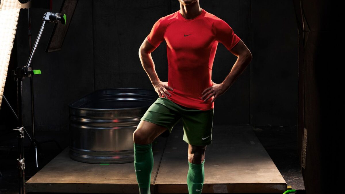 Athlete in a red shirt and green shorts stands with hands on hips in a photography studio; a soccer ball at his feet.