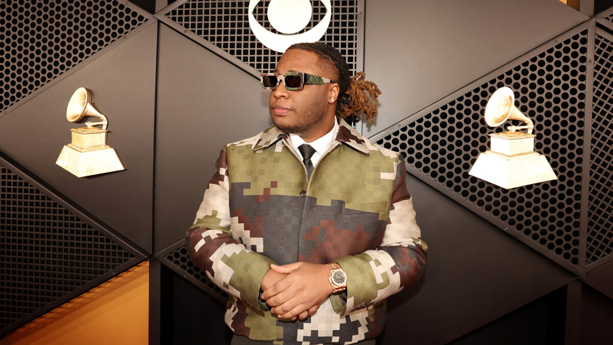 Man in camo-patterned suit with folded arms stands against geometric wall, wearing sunglasses and a gold watch, with vintage gramophone decor in the background.