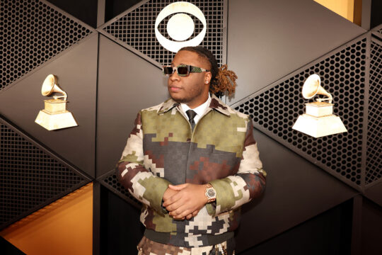 Man in camo-patterned suit with folded arms stands against geometric wall, wearing sunglasses and a gold watch, with vintage gramophone decor in the background.