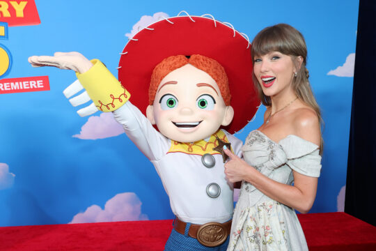 Woman in a light floral dress posing with a life-size Woody mascot on a blue sky backdrop with clouds and a red carpet detailing her arm around the character's shoulder.