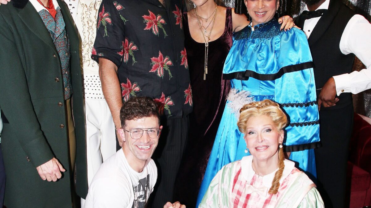Group of six adults in vintage costumes posing together in front of a sparkly backdrop at a party.