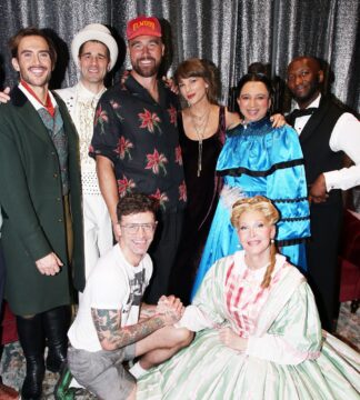 Group of six adults in vintage costumes posing together in front of a sparkly backdrop at a party.