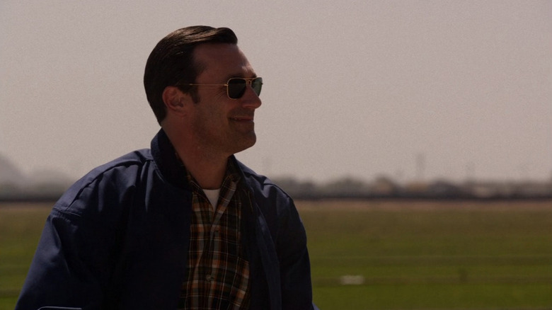 Jon Hamm as Don Draper sitting with sunglasses in front of a green field in Mad Men
