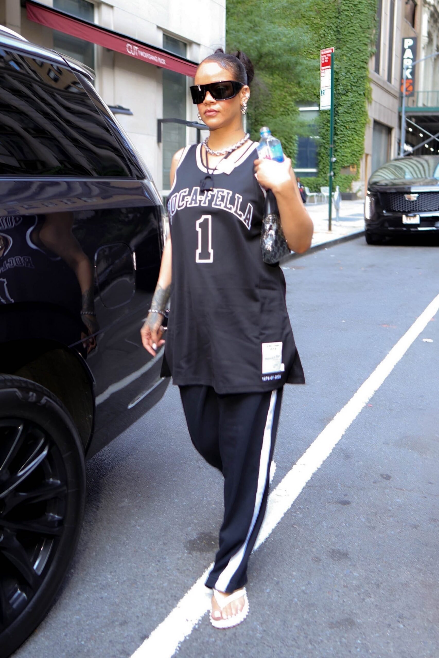 A person wearing a sports jersey and pants walks beside a black vehicle in an urban setting.