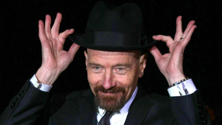 Bryan Cranston with a goatee while wearing a black suit and tie with a black trilby. He has his hands on the hat as if he's repositioning it on his head.