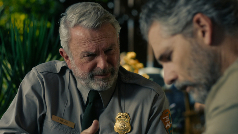 On the left is Sam Neill with gray hair and a beard wearing a Park Ranger Chief uniform, on the right is Eric Bana with graying black hair wearing a khaki shirt — Bana is out of focus.