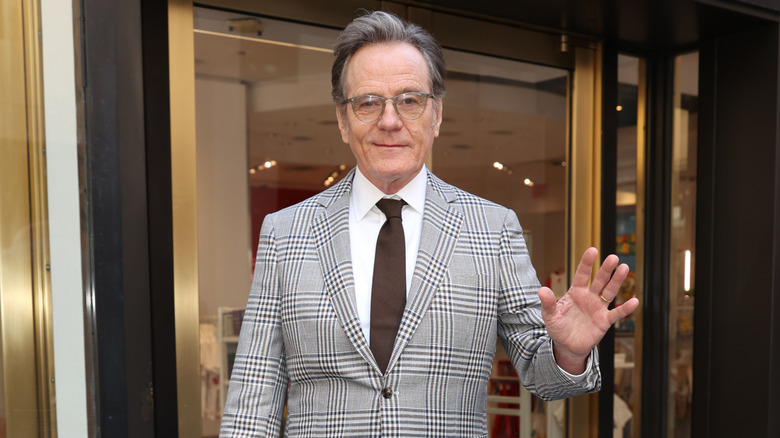 Bryan Cranston with graying hair wearing silver glasses and a gray checked suit jacket with a white shirt and a brown tie.
