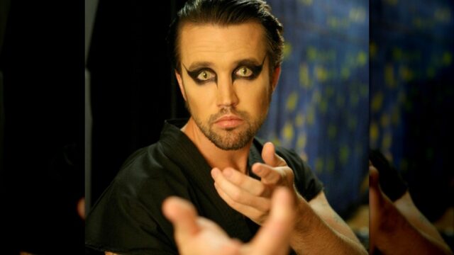 A man with heavy black eye makeup reaches toward the camera against a dark, patterned background.