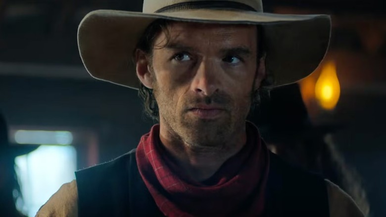 A man in a wide-brimmed hat and red neckerchief looks aside in a dimly lit interior.