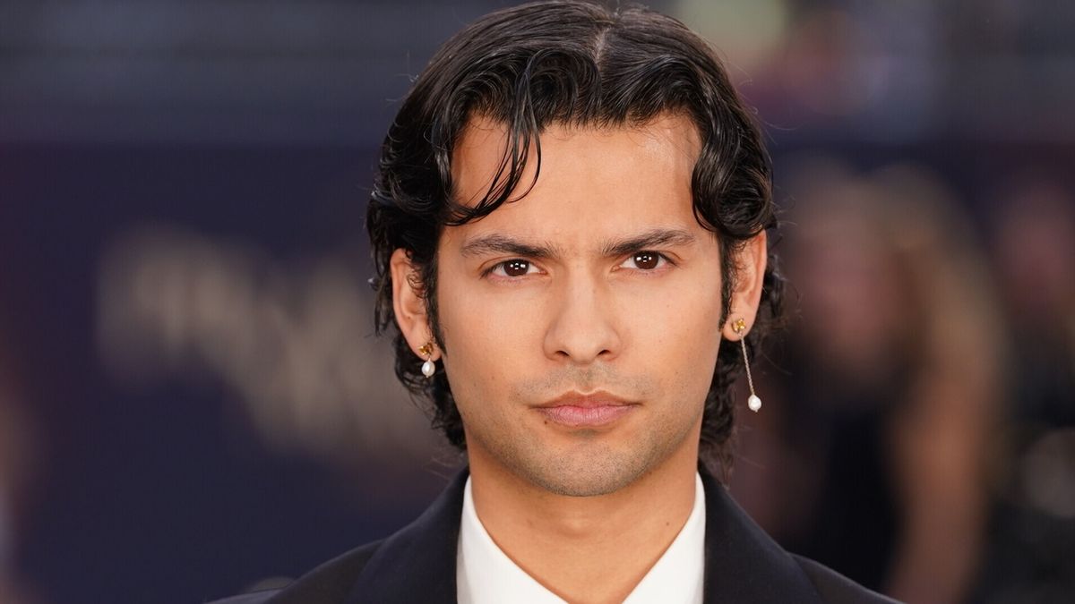 Close-up portrait of a man with wavy dark hair, dangling earrings, and a black suit against a blurred background.