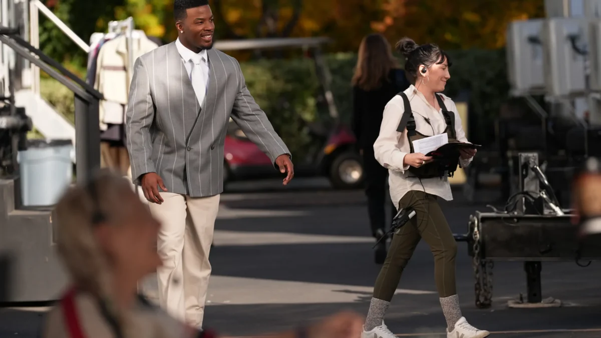 A smiling man in a gray striped suit walks past a crew member holding papers outdoors.
