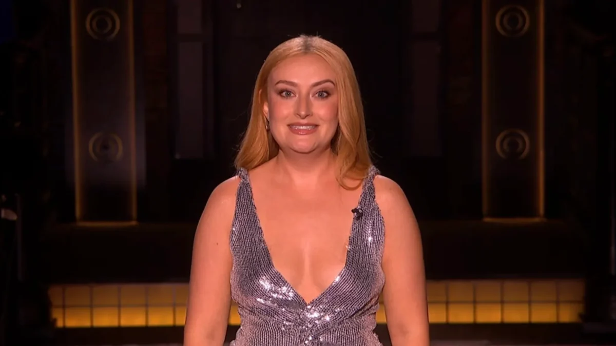 Blonde woman in a shimmering silver sleeveless dress smiles on a dimly lit stage.