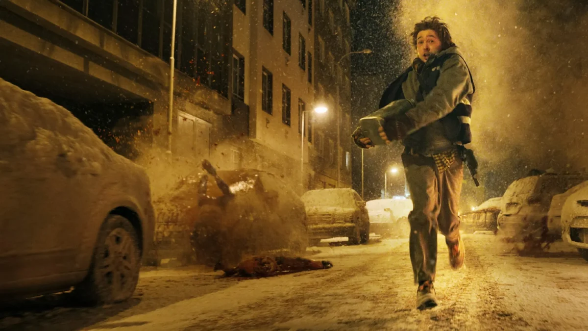 A person runs through a snowy, debris-filled street carrying a bundled child as cars and buildings line the road.