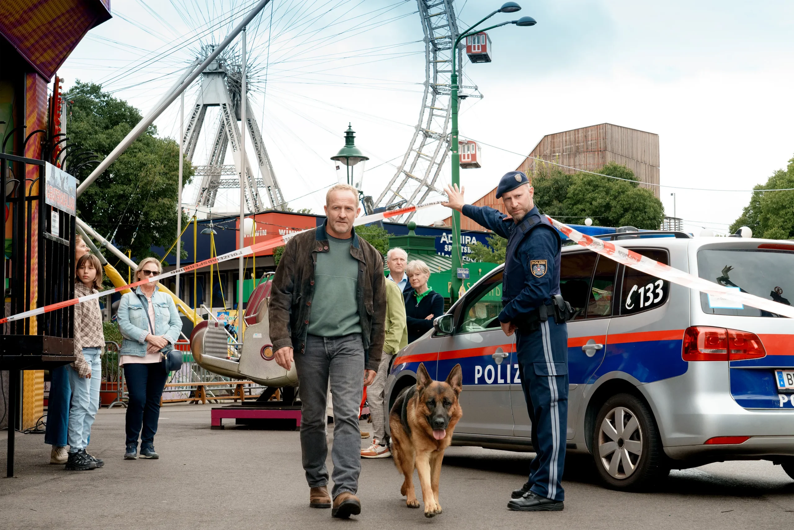 A man walks a dog past a police car and barrier tape near a Ferris wheel.