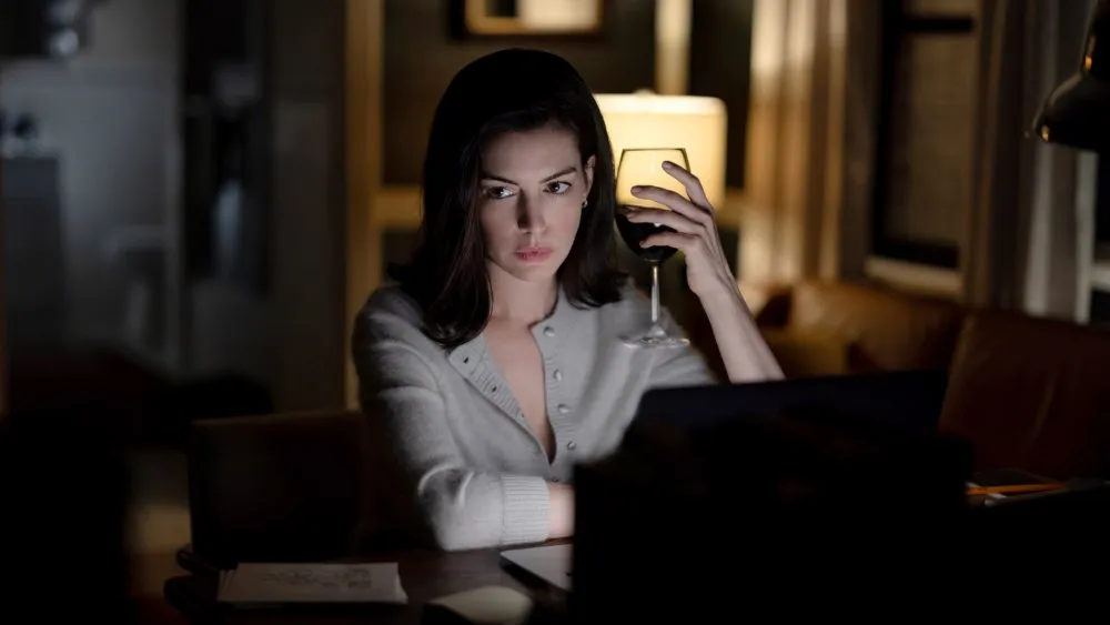 A woman in a dim room holds a wine glass while looking at a laptop.