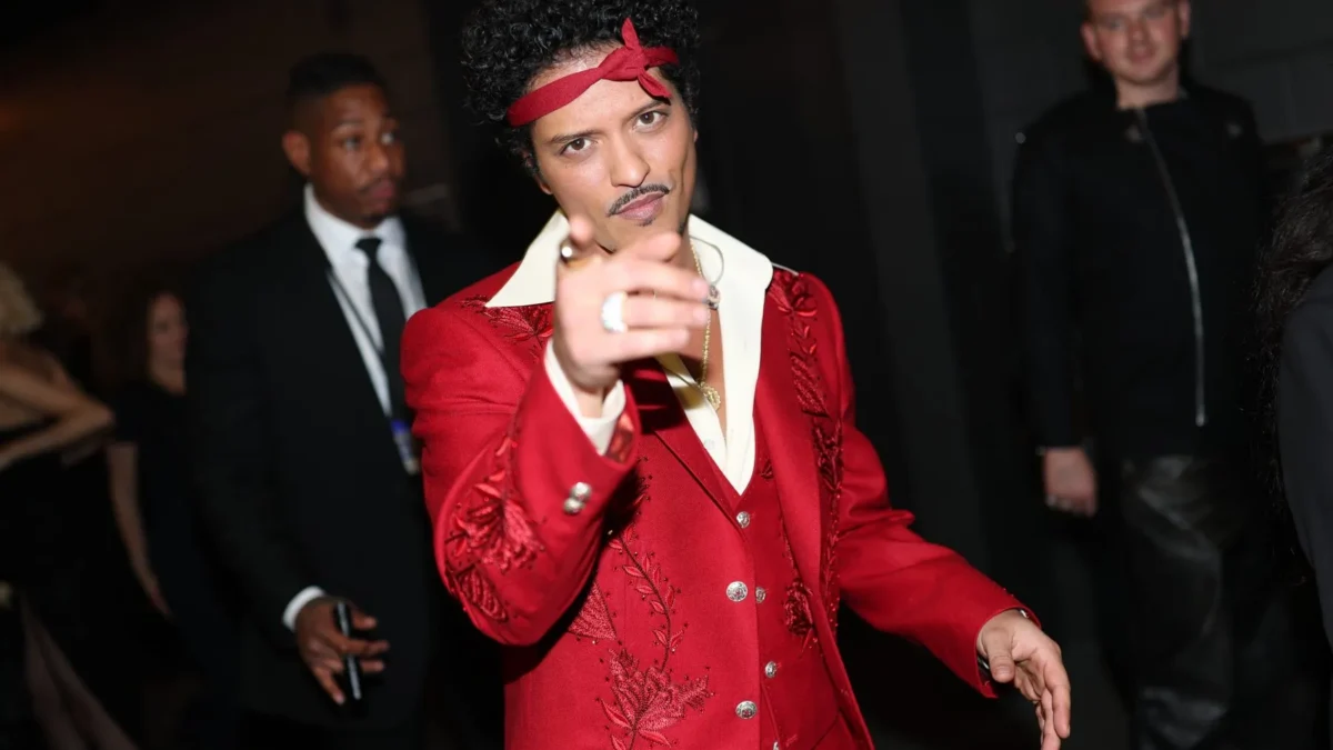 A man in a red embroidered suit and headband points toward the camera amid people in a dark setting.