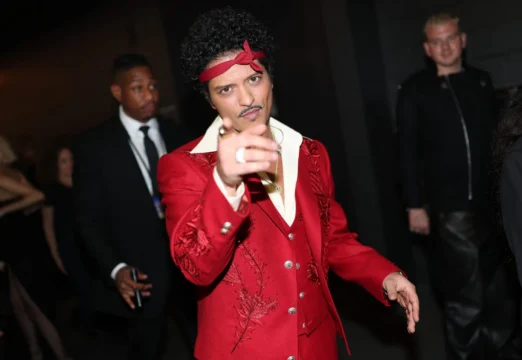A man in a red embroidered suit and headband points toward the camera amid people in a dark setting.