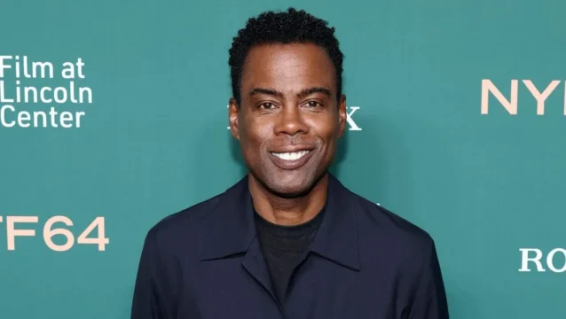 A smiling man in a dark jacket stands before a teal backdrop with white event lettering.