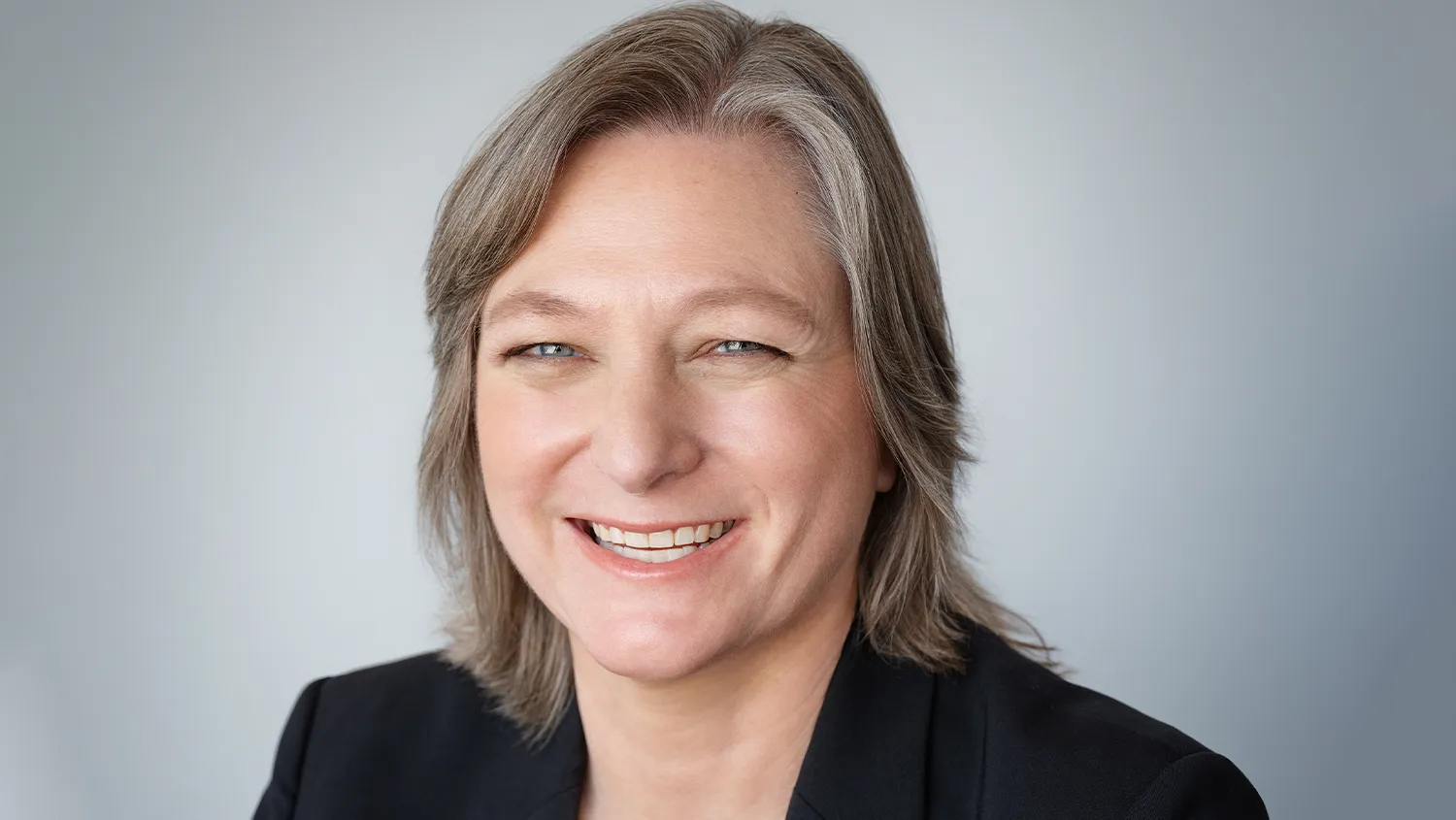 Smiling person with gray hair wearing a black blazer against a light gray background.