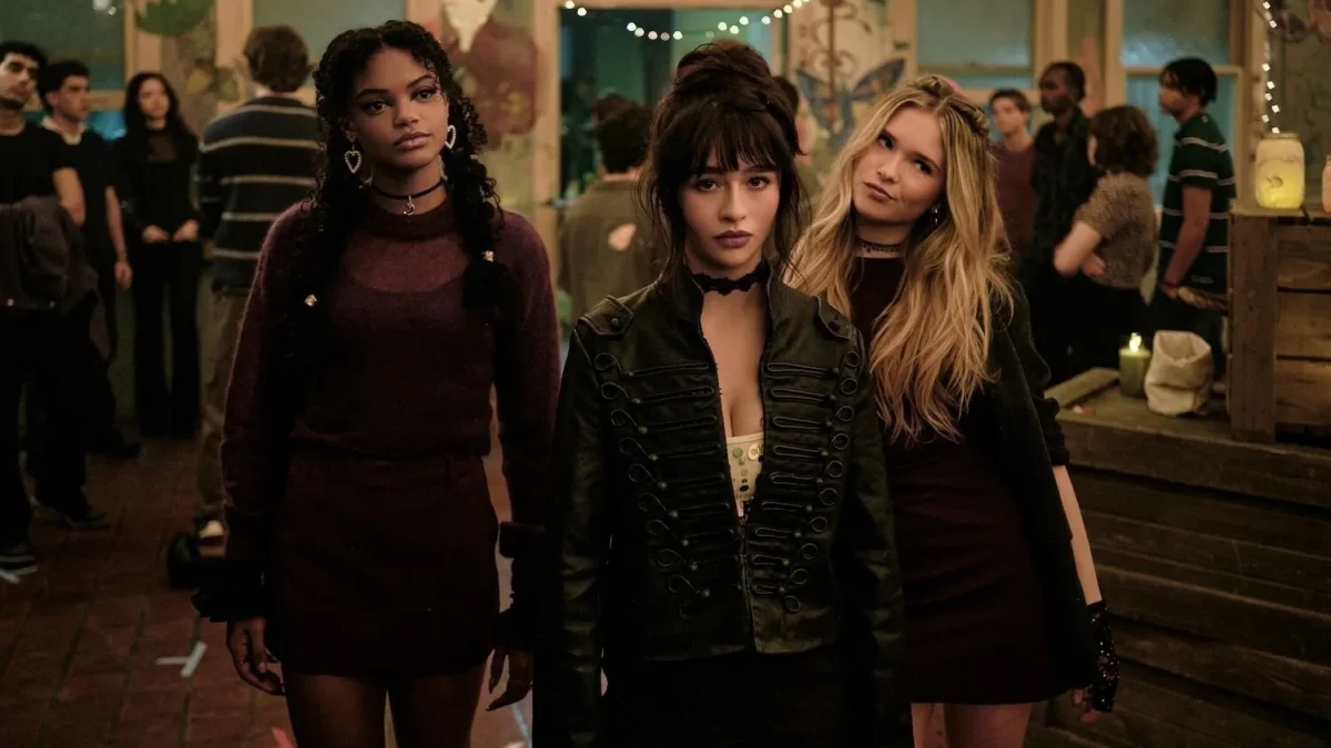 Three young women stand together in a dimly lit, crowded room decorated with string lights.