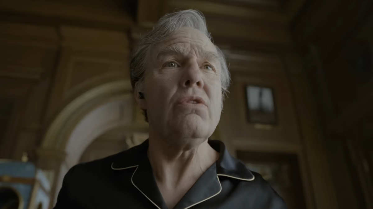 An older man in a black collared shirt looks upward in a dim, ornate room.