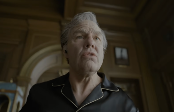 An older man in a black collared shirt looks upward in a dim, ornate room.