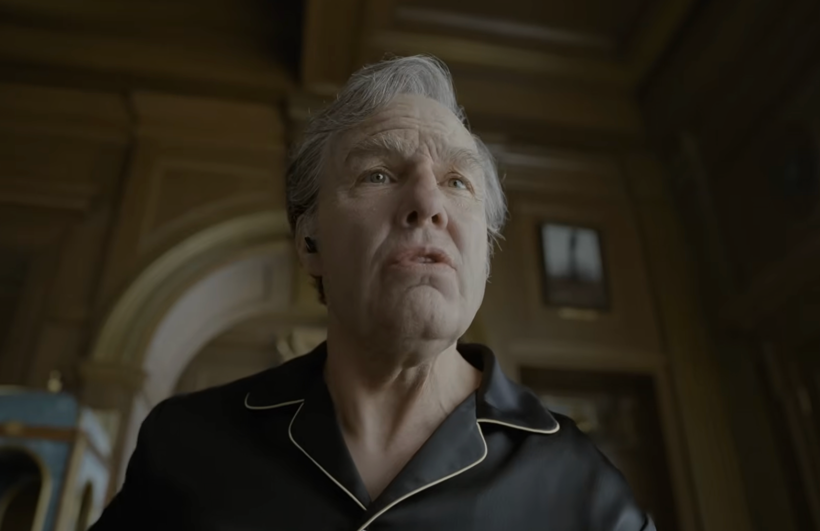 An older man in a black collared shirt looks upward in a dim, ornate room.