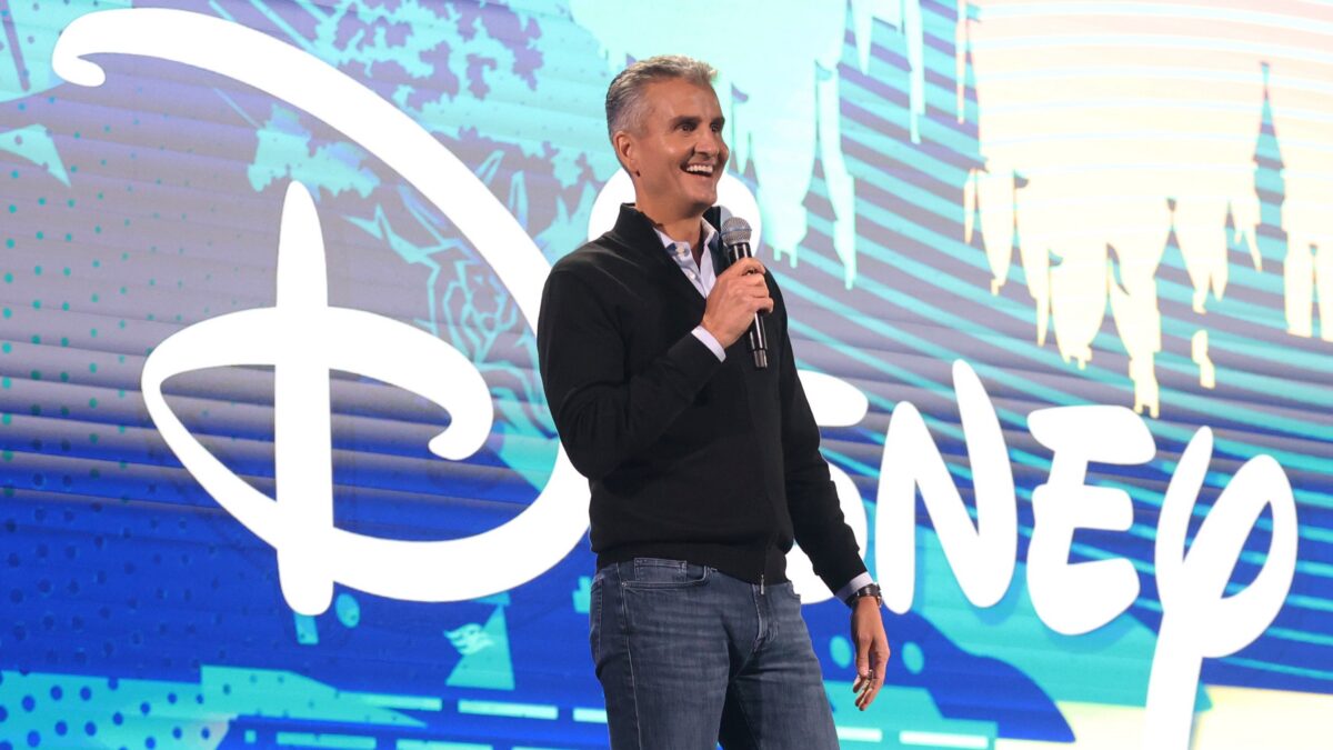A man holds a microphone onstage in front of a large Disney logo and colorful graphic backdrop.