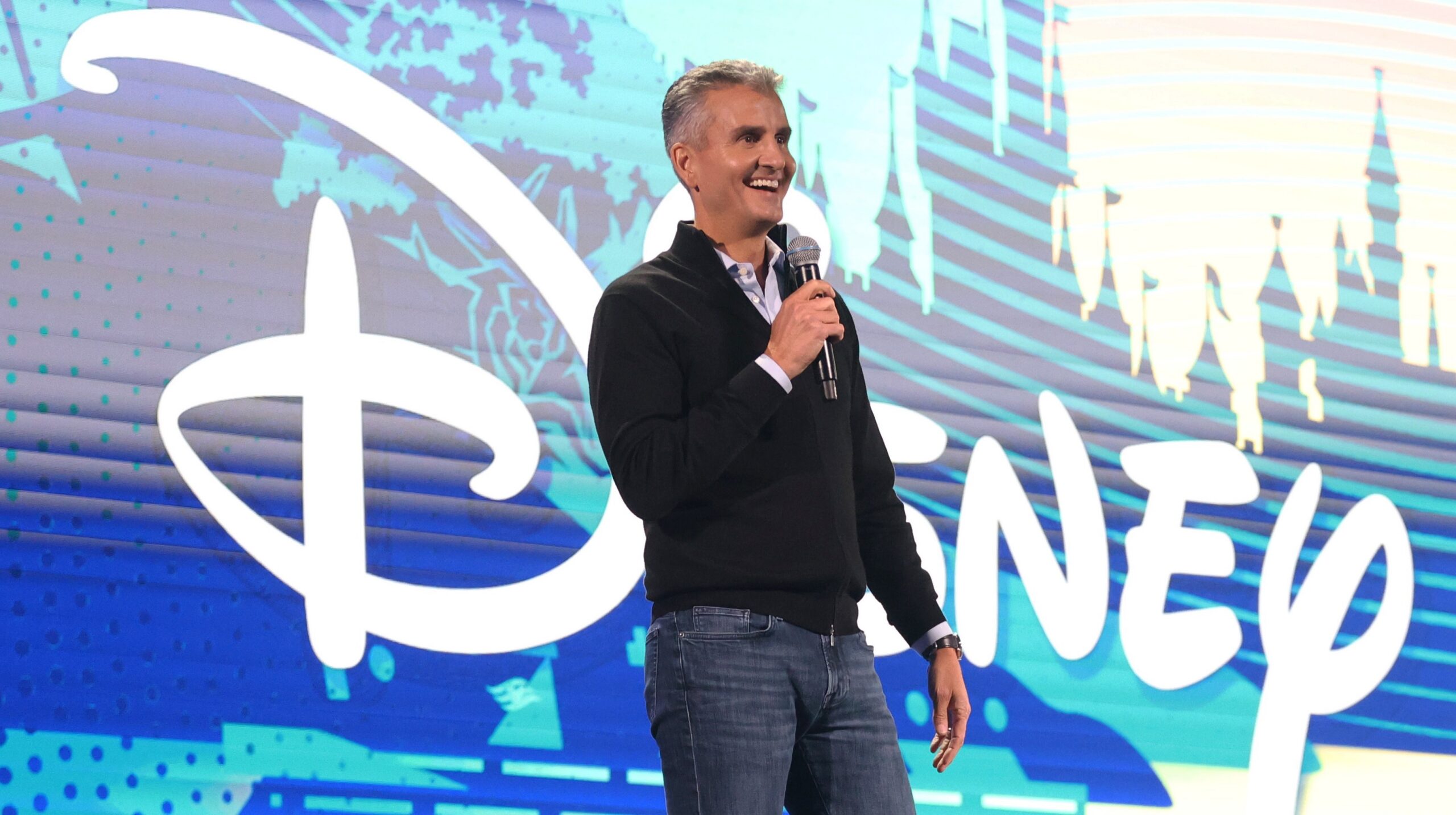 A man holds a microphone onstage in front of a large Disney logo and colorful graphic backdrop.