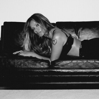 A long-haired person lies on a dark couch, looking toward the camera, with a heart tattoo on one arm.