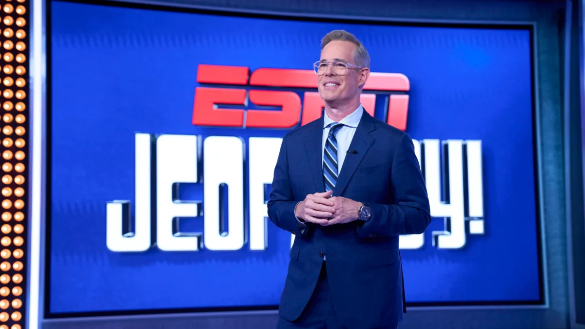 A smiling man in a suit stands before a blue ESPN Jeopardy! backdrop.