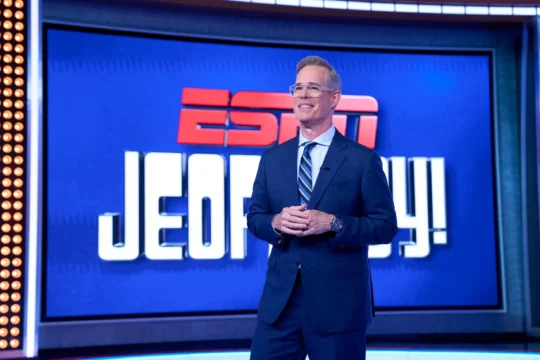 A smiling man in a suit stands before a blue ESPN Jeopardy! backdrop.