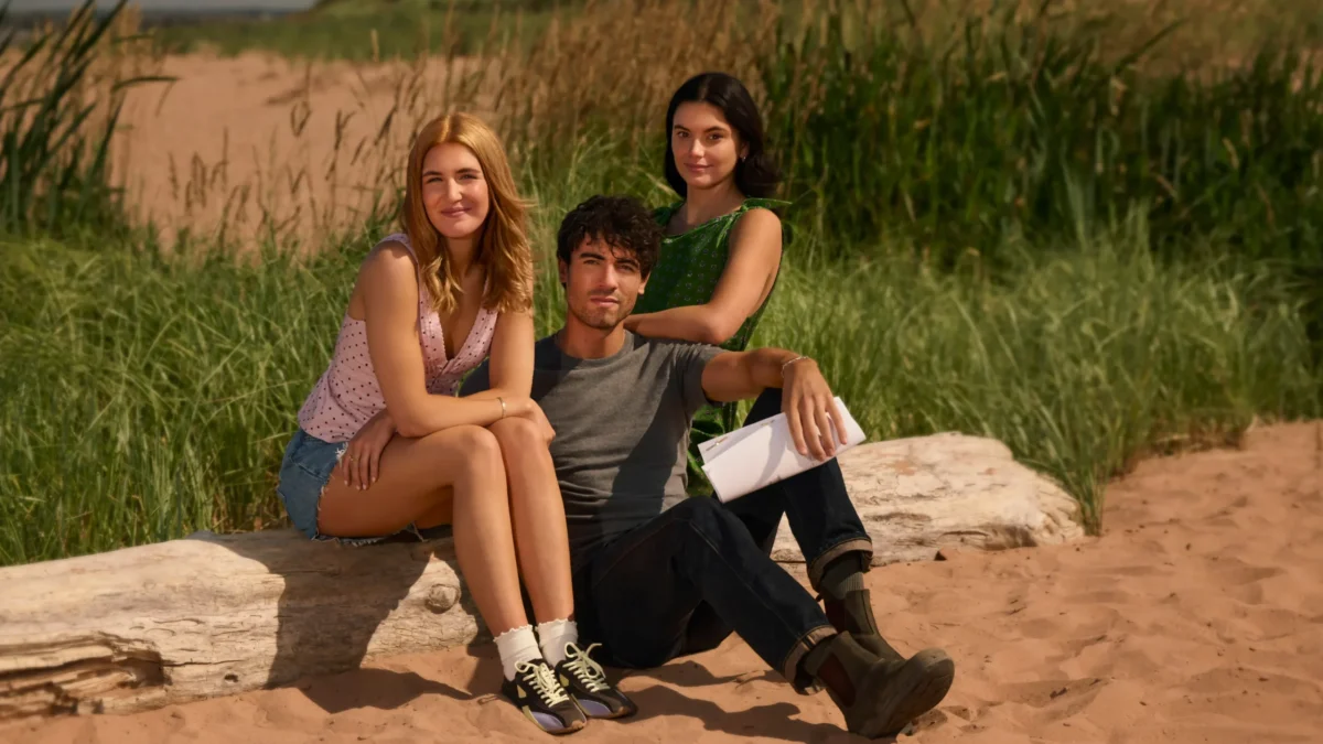 "Three young adults sitting on a log at a sandy beach with tall grass in the background, casual summer clothing, one holding a script, relaxed atmosphere in a natural outdoor setting."