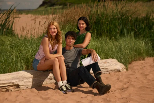 "Three young adults sitting on a log at a sandy beach with tall grass in the background, casual summer clothing, one holding a script, relaxed atmosphere in a natural outdoor setting."