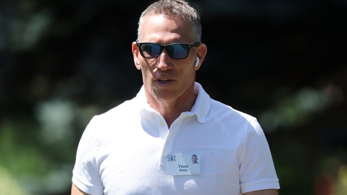 A man in sunglasses and a white polo shirt wears an earbud and name badge outdoors.