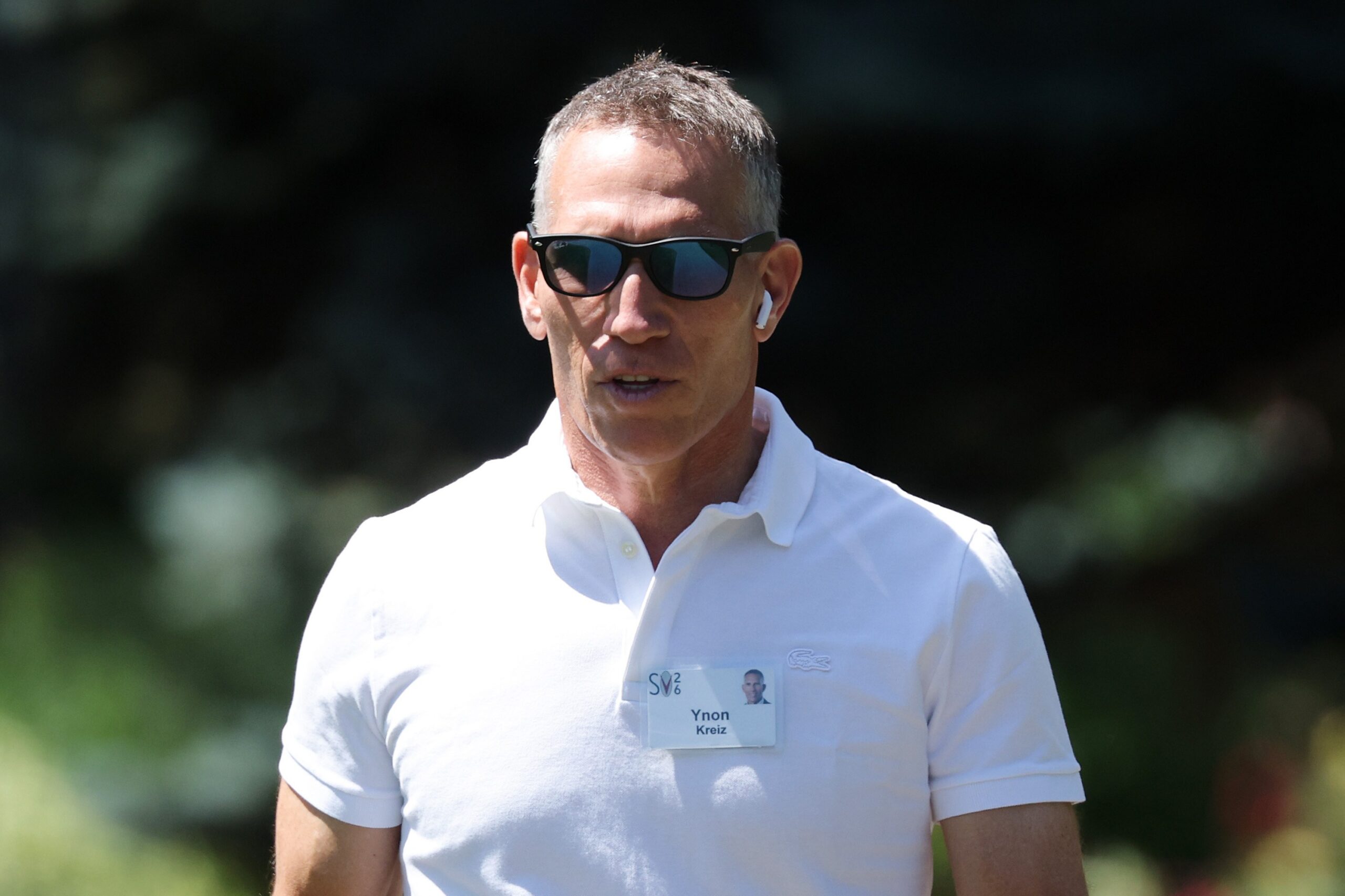 A man in sunglasses and a white polo shirt wears an earbud and name badge outdoors.