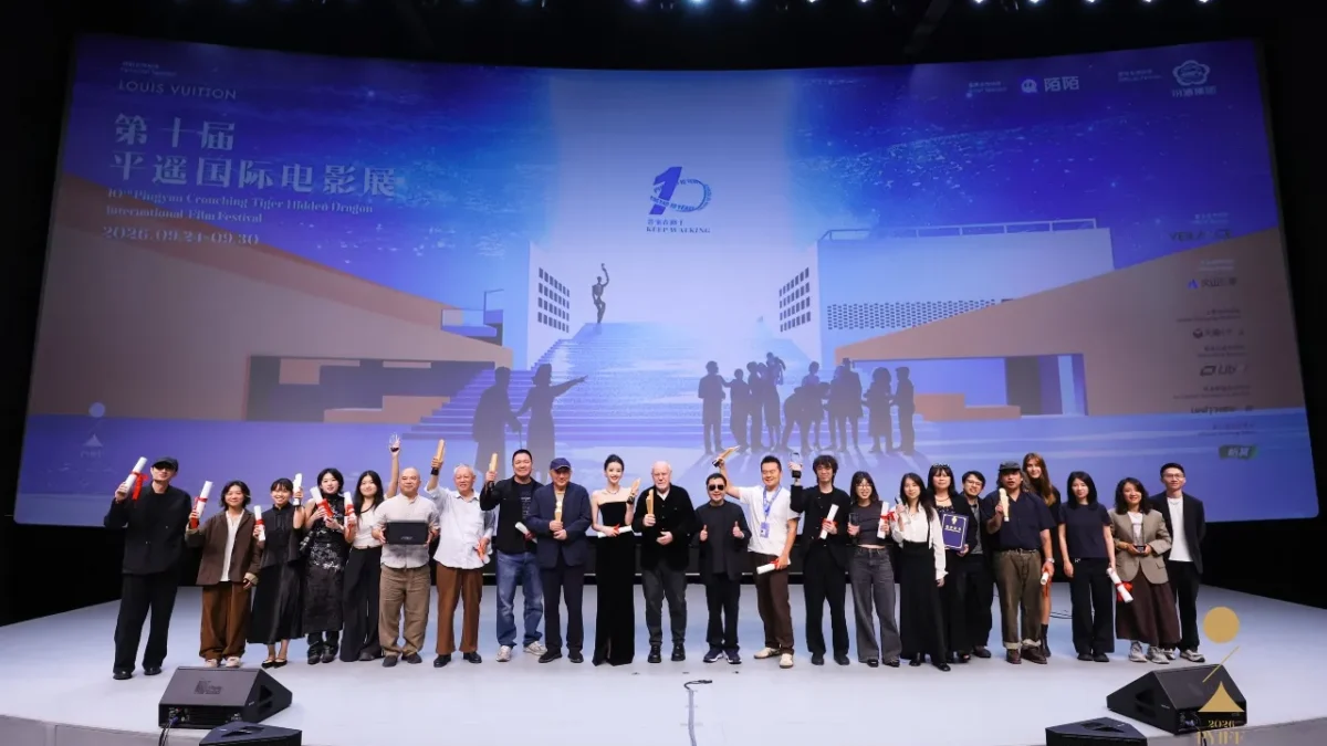A large group poses onstage holding awards and scrolls before a brightly lit festival backdrop.
