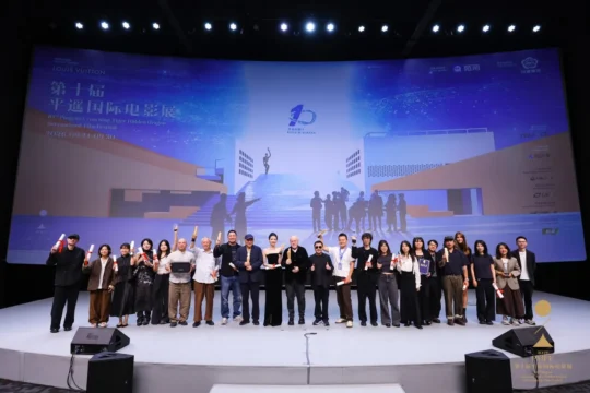 A large group poses onstage holding awards and scrolls before a brightly lit festival backdrop.