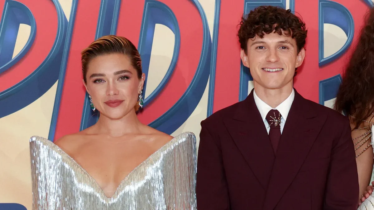 Two people pose before a colorful lettered backdrop, one in a silver dress and one in a burgundy suit.