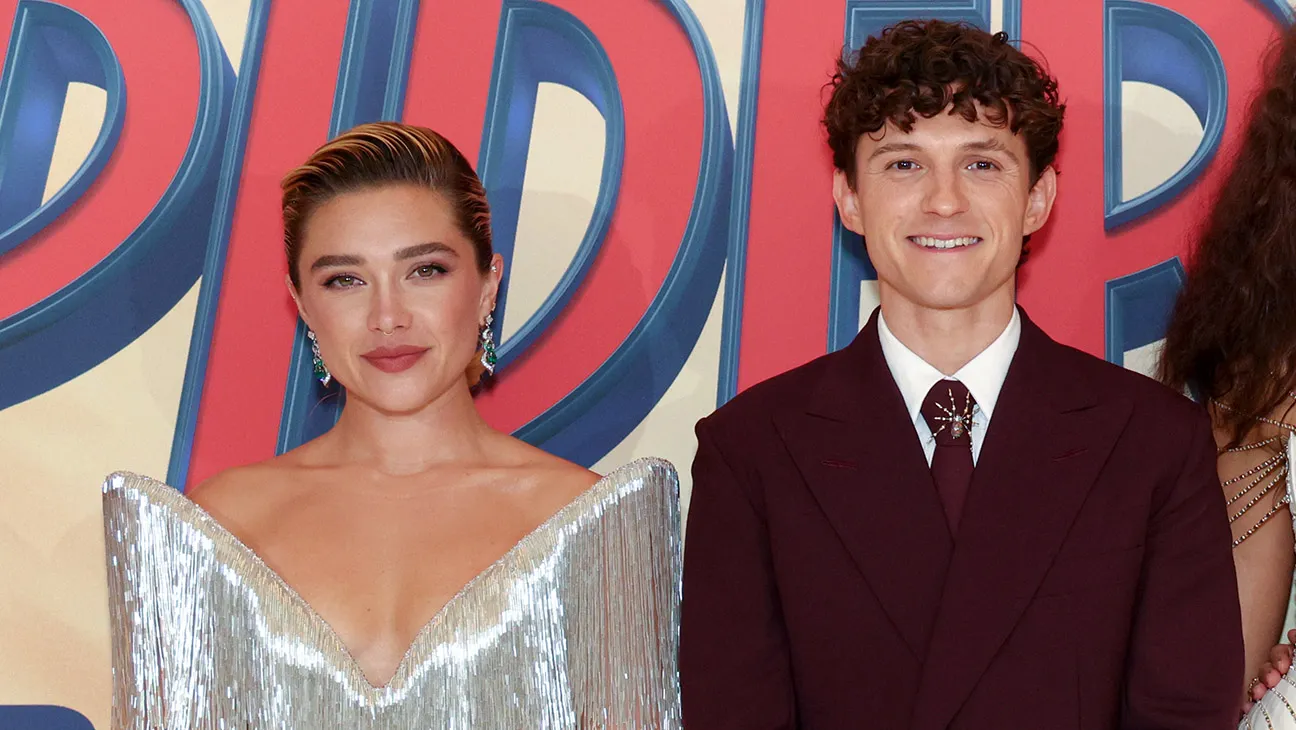 Two people pose before a colorful lettered backdrop, one in a silver dress and one in a burgundy suit.