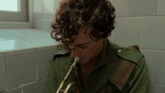 A curly-haired person in a green shirt plays a trumpet beside tiled walls.