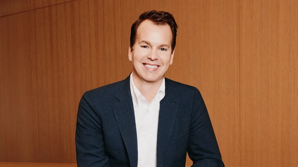 A smiling man in a navy blazer and white shirt poses against a wood-paneled wall.