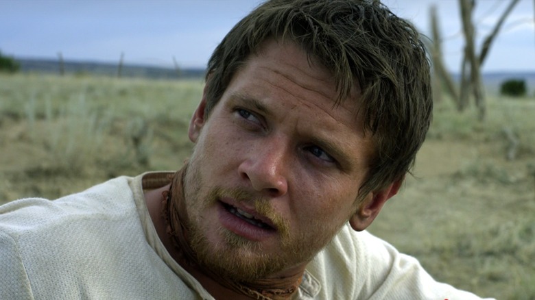 A close-up of a bearded man in a light shirt looking sideways in a dry, open landscape.